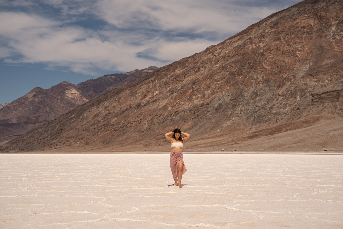 Melissa with both hands raised to her hair, standing on white salt beneath mountains and streaked clouds.