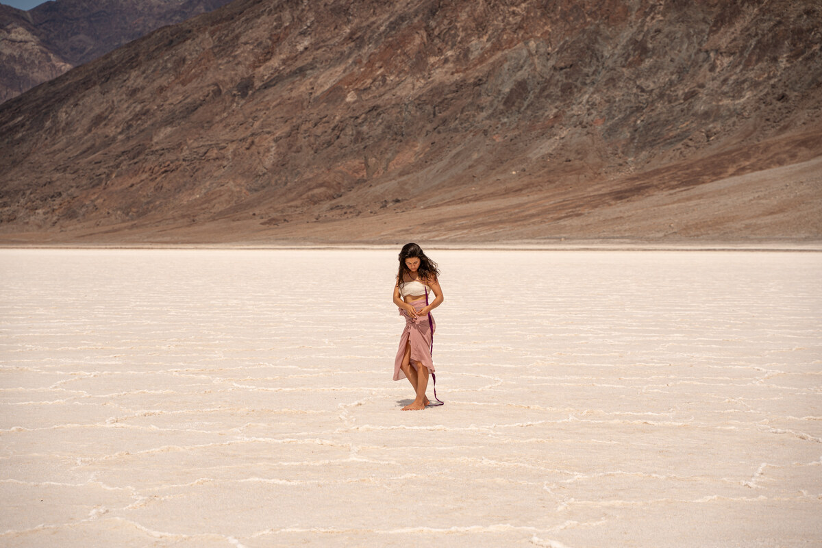 Melissa looking downward, her hands near her waist, surrounded by pale salt and a brown mountain slope.